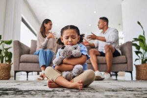 a child hugs a teddy bear and sit on the floor while their parents argue on the couch behind them showing the impact of opioid addiction